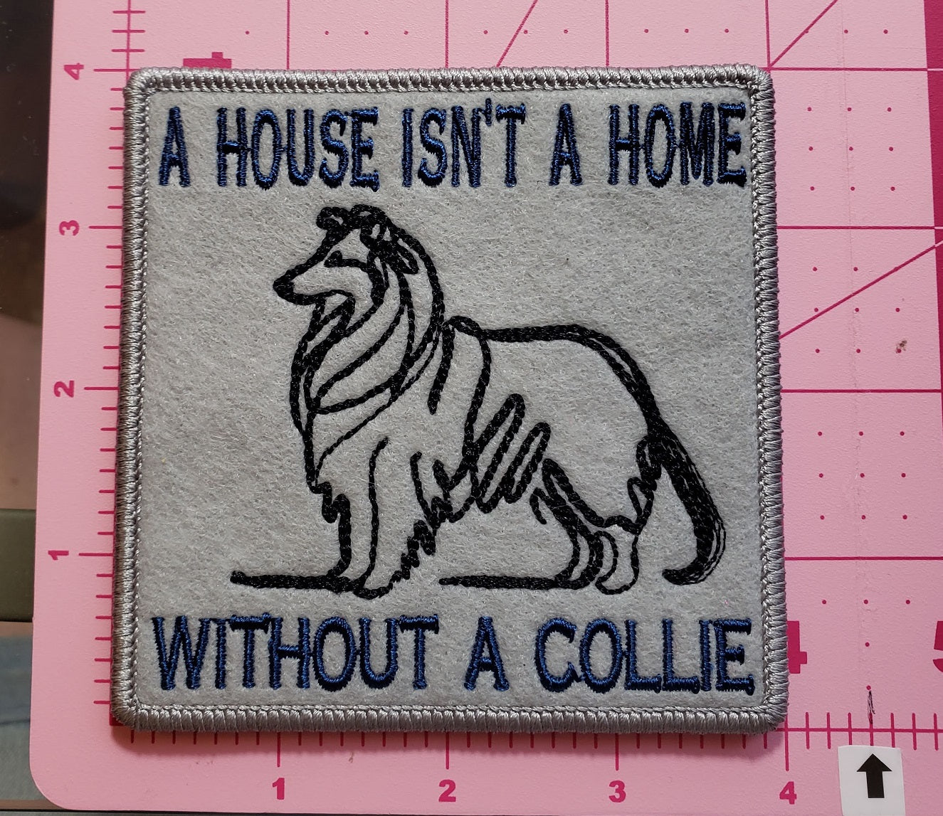 A Gray Felt Mug Rug with a line drawing a Collie with the text " A house isn't a home without a collie" with a high luster sterling border on a pink cutting mat showing the size of the item