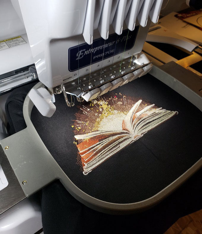Book of Wonder image being stitched on a black cotton tote on multi-needle embroidery machine
